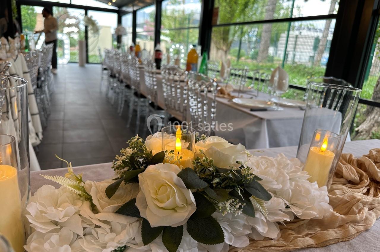 Centre de table floral avec roses blanches et bougies LED, dans une salle de réception lumineuse avec tables dressées.