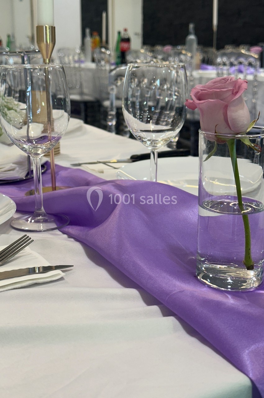 Table dressée avec nappe blanche, chemin de table violet, verre contenant une rose rose et verres à vin vides.