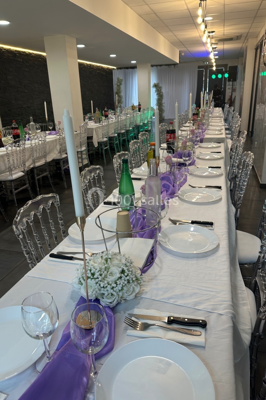 Grande table de réception dressée avec nappes blanches, chaises transparentes, bougies et décorations violettes.