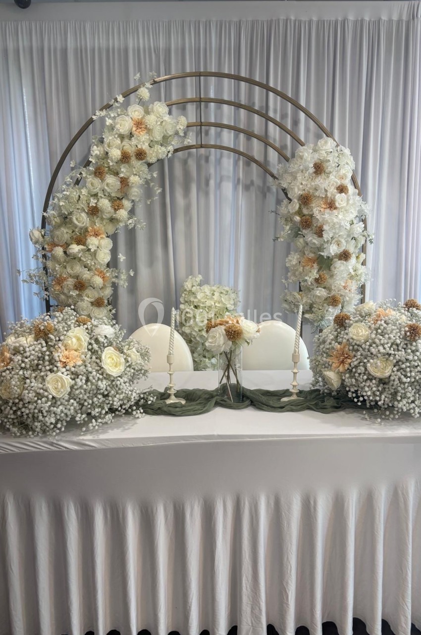 Table décorée pour un mariage avec fleurs blanches et beige, bougies et arche florale en arrière-plan.
