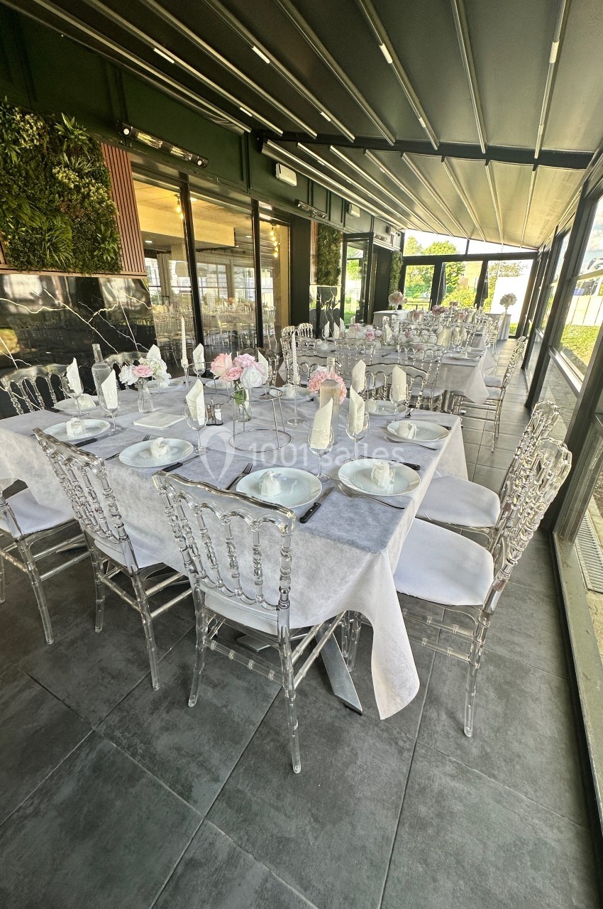 Salle de réception avec tables dressées, nappes blanches, chaises transparentes et décoration florale.