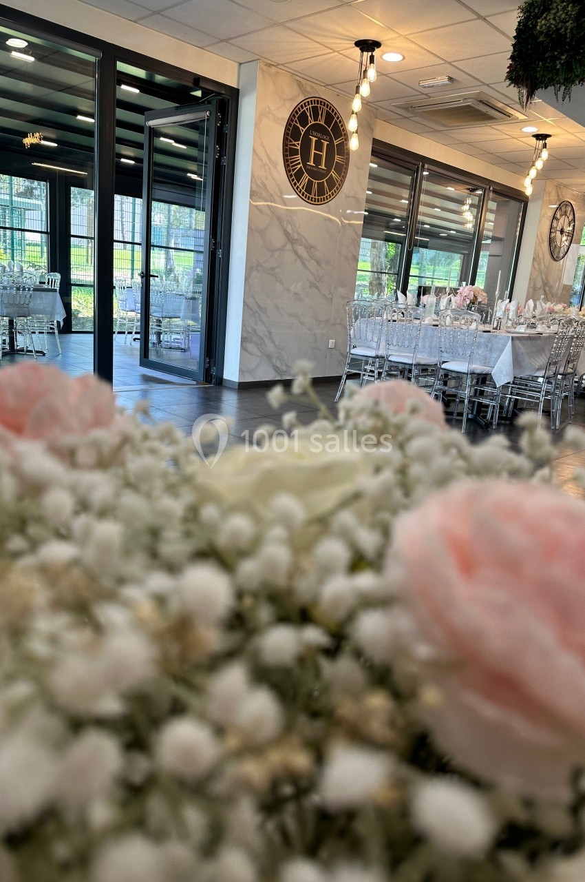 Salle de réception élégante avec tables dressées, vue partielle sur des fleurs en premier plan et grandes baies vitrées.