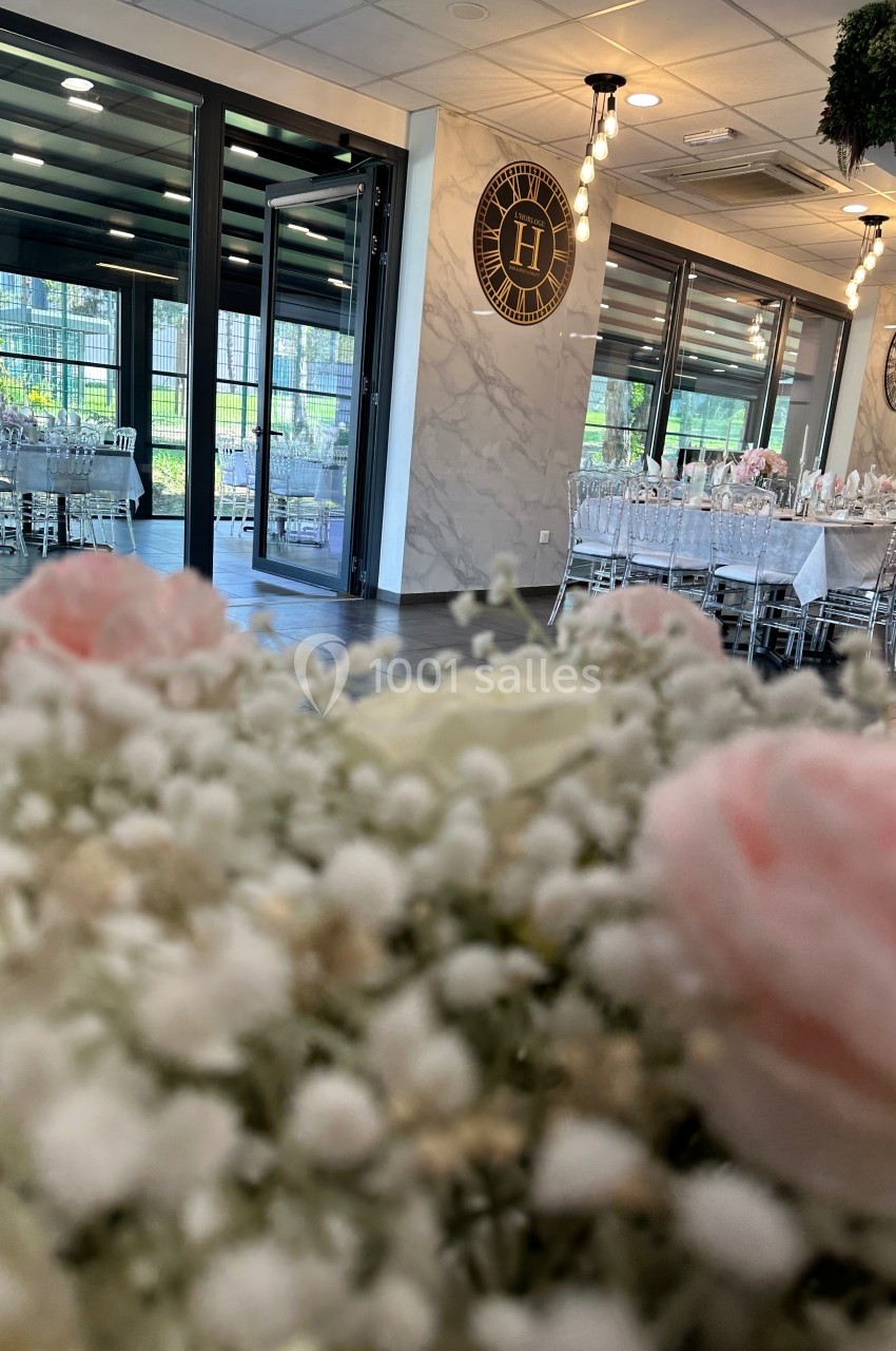 Salle de réception lumineuse avec tables dressées, chaises transparentes et décor floral en premier plan.