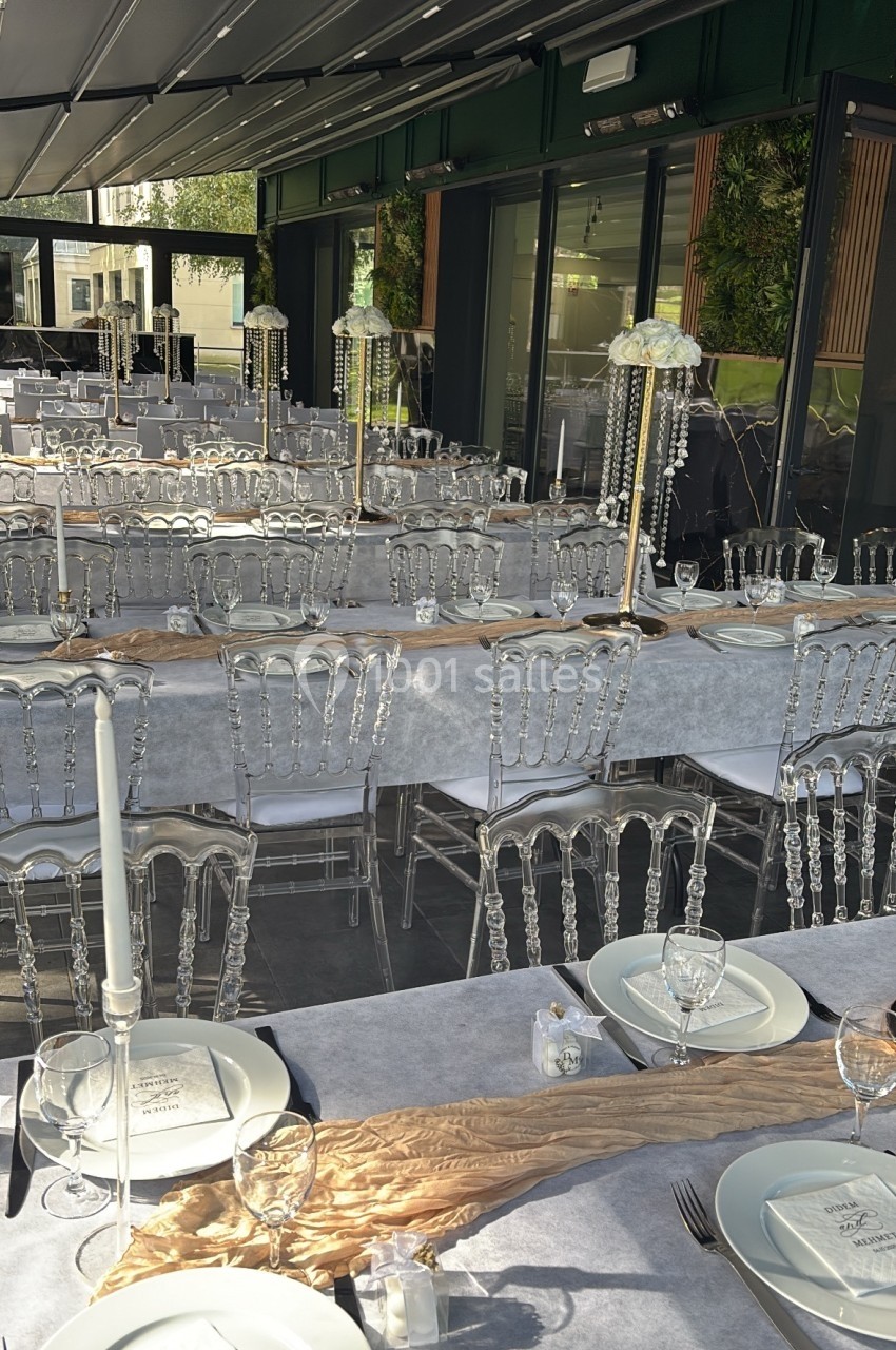 Salle de réception élégamment dressée avec des tables alignées, nappes grises, chaises transparentes et décorations dorées.