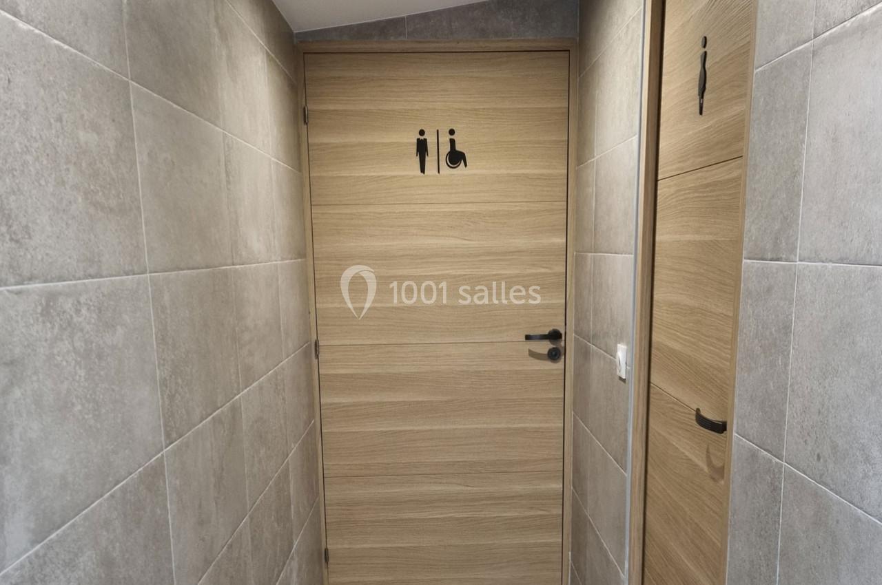 Porte de toilettes accessibles avec pictogrammes homme, femme et fauteuil roulant, entourée de murs carrelés gris.