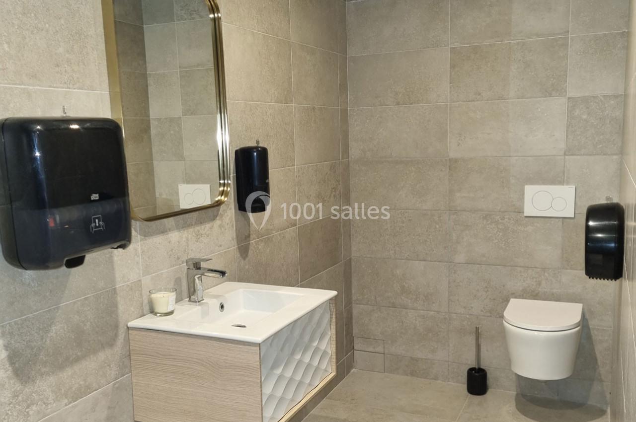 Salle de bain moderne avec lavabo, miroir doré, distributeurs de savon, toilettes suspendues et carrelage beige.