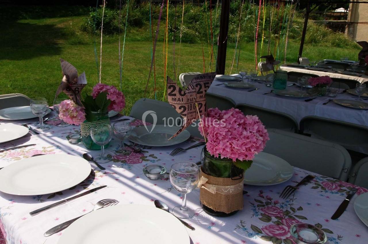 Table dressée en extérieur avec nappes fleuries, assiettes blanches, bouquets d'hortensias roses et décorations en papier.