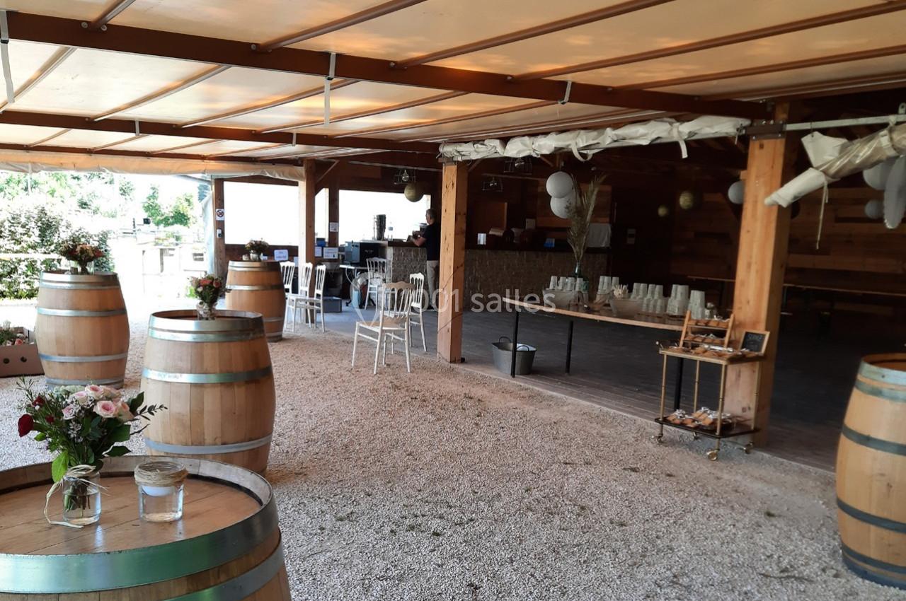 Salle de réception en plein air avec tonneaux en bois, tables décorées et espace couvert en arrière-plan.