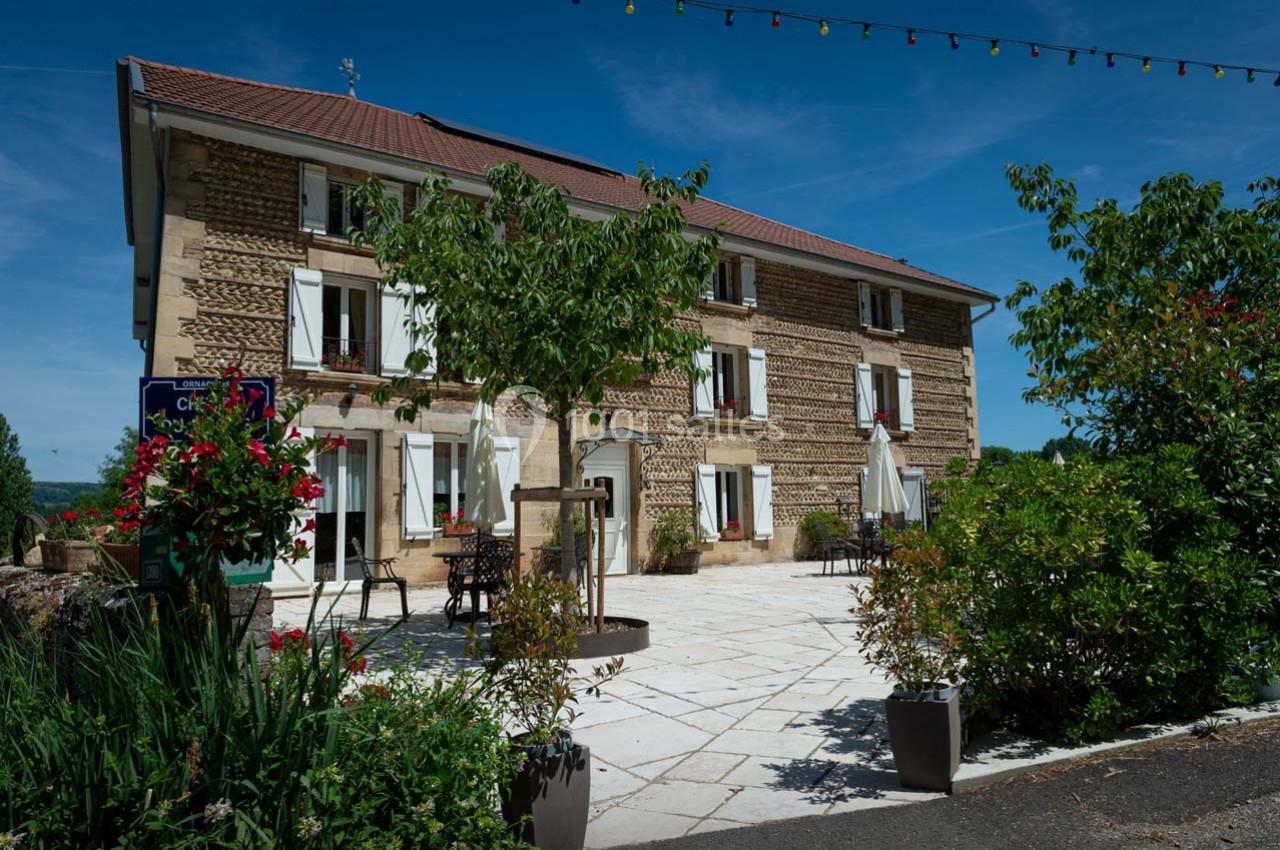 Façade d'une maison en pierre avec terrasse fleurie, arbres et ciel bleu en arrière-plan.