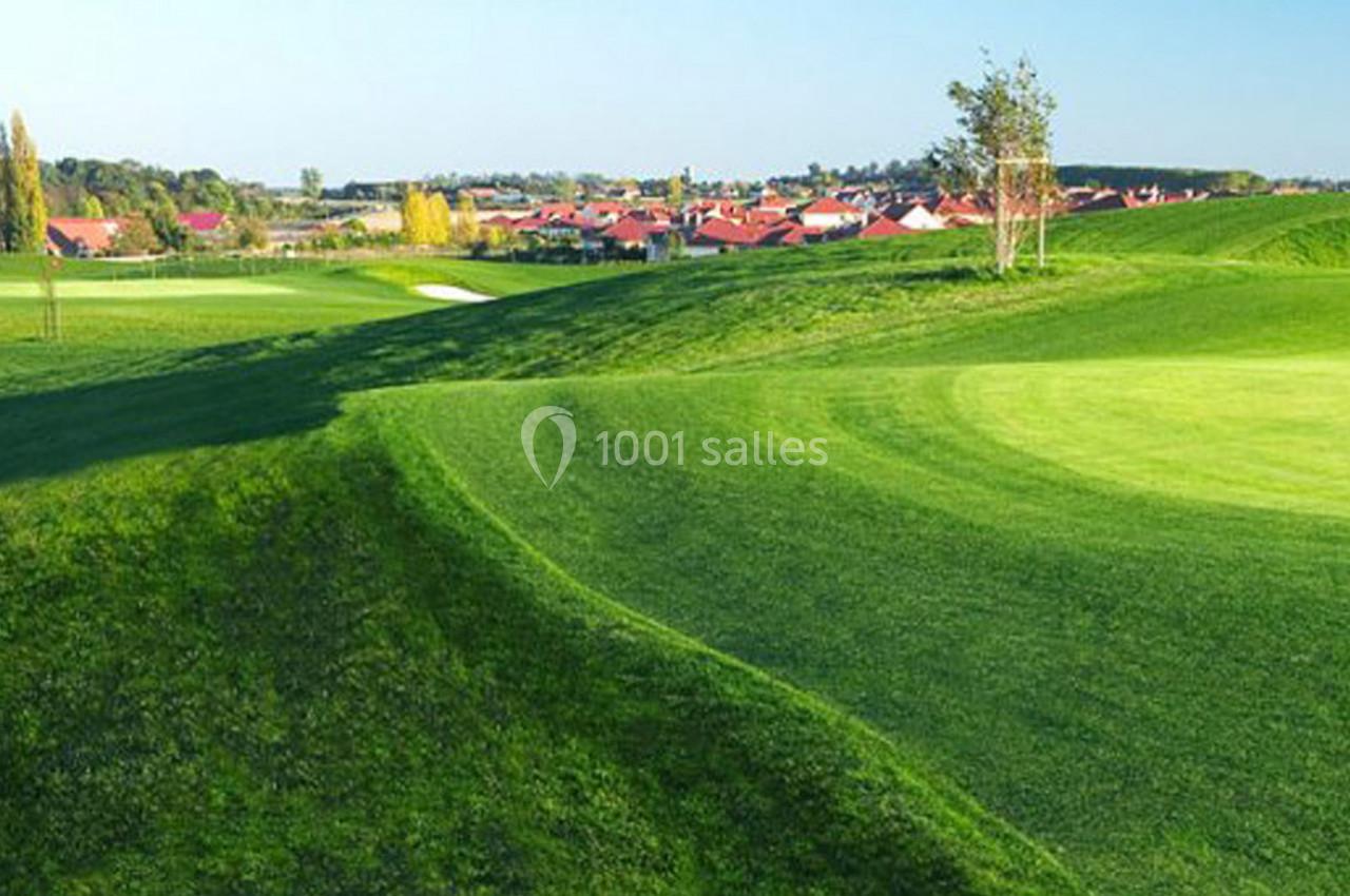 Paysage de golf avec pelouses verdoyantes, reliefs ondulés et maisons en arrière-plan sous un ciel dégagé.