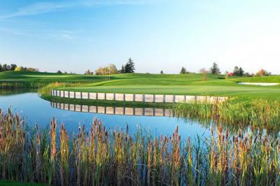 Paysage de golf avec pelouses verdoyantes, reliefs ondulés et maisons en arrière-plan sous un ciel dégagé.