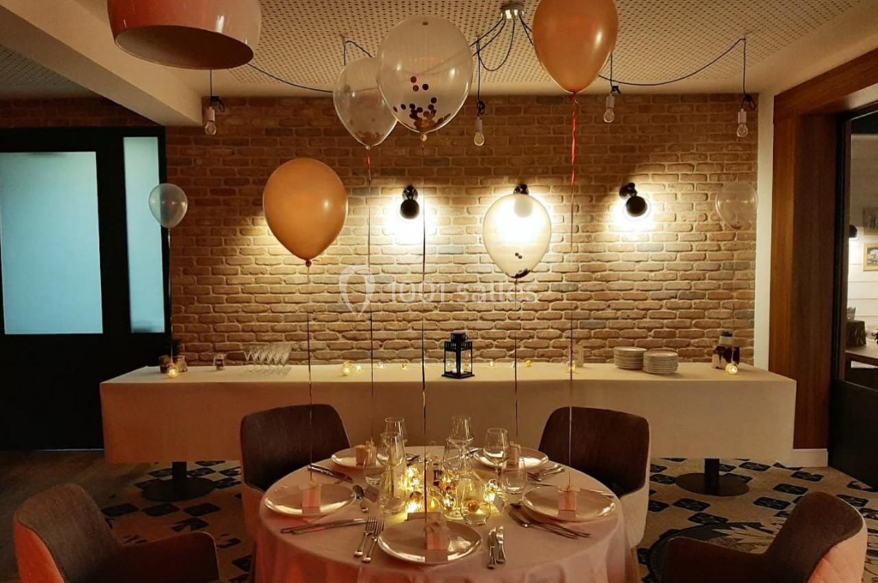 Salle décorée pour une réception, avec ballons suspendus, table dressée et éclairage tamisé.