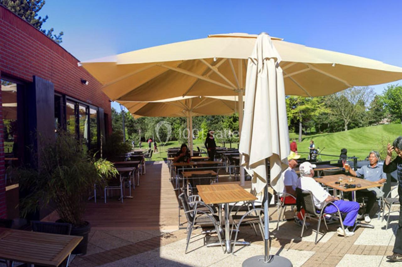 Terrasse extérieure d'un café avec des tables, des chaises et des parasols, entourée de verdure et de clients assis.