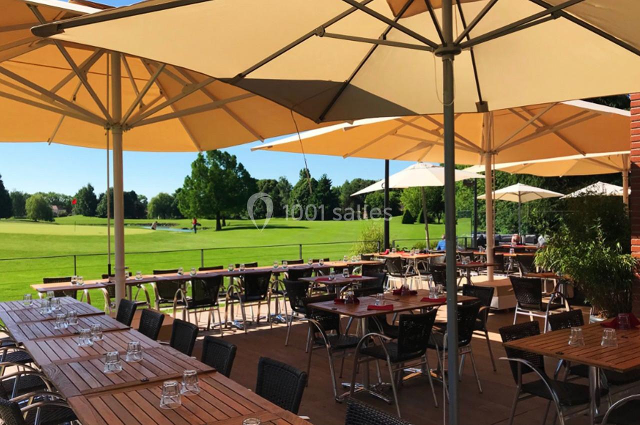 Terrasse ensoleillée avec tables en bois, chaises et parasols, donnant sur un terrain de golf verdoyant.