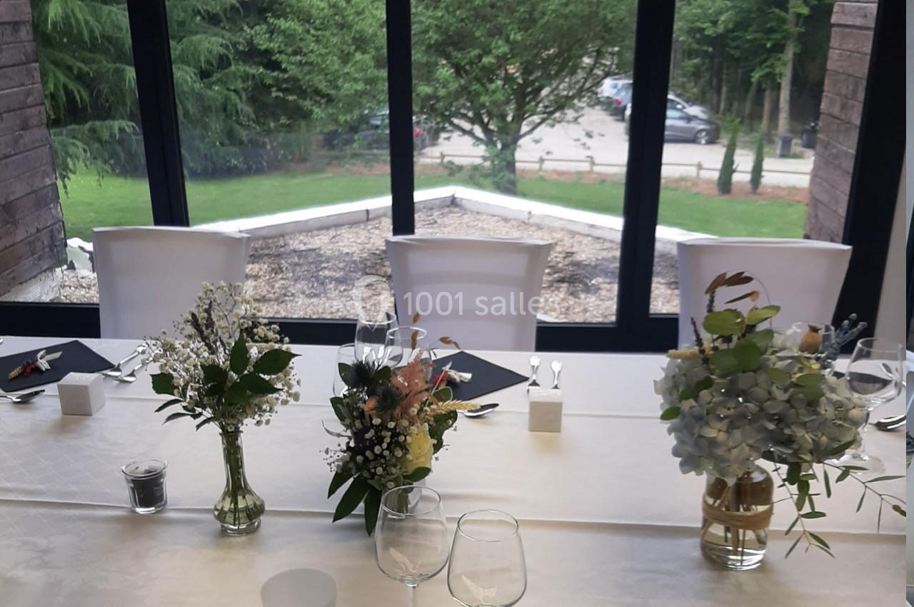 Table dressée avec nappes blanches, décorée de bouquets de fleurs, près d'une baie vitrée donnant sur un espace arboré.