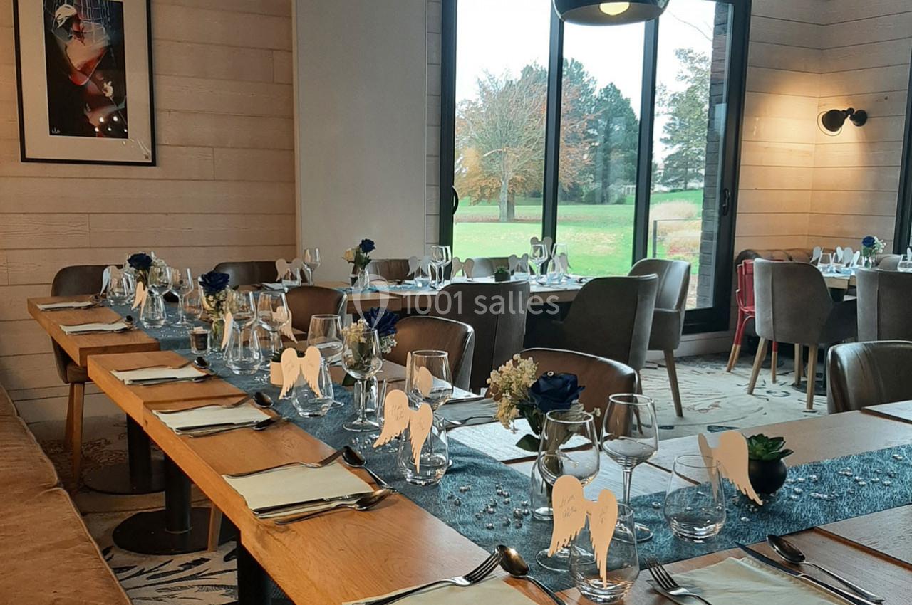 Salle de restaurant décorée pour un événement, avec tables dressées, serviettes pliées et vue sur un jardin extérieur.