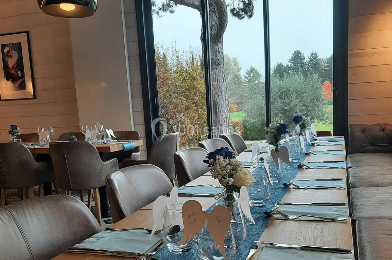 Salle de restaurant lumineuse avec une longue table dressée, décorée de fleurs et vue sur un jardin arboré.