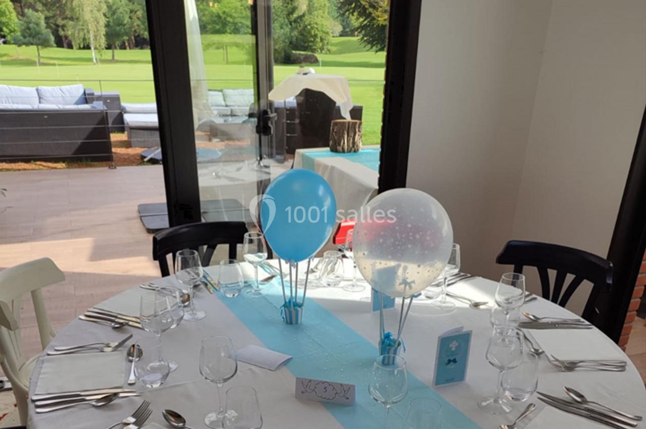 Table ronde dressée pour un événement, décorée de ballons bleus et blancs, avec vue sur un jardin verdoyant.