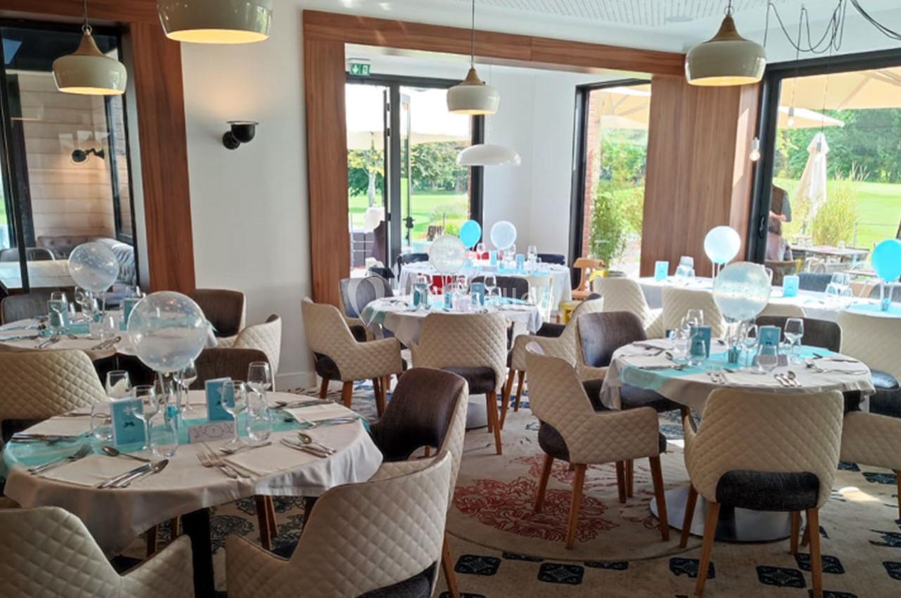 Salle de réception lumineuse avec tables rondes dressées, décorées de ballons transparents et vue sur un jardin.