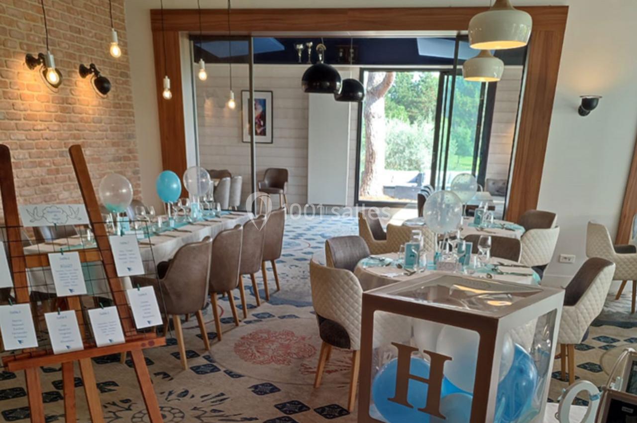 Salle de réception décorée avec des tables dressées, des ballons bleus et blancs, et un chevalet avec des cartes.