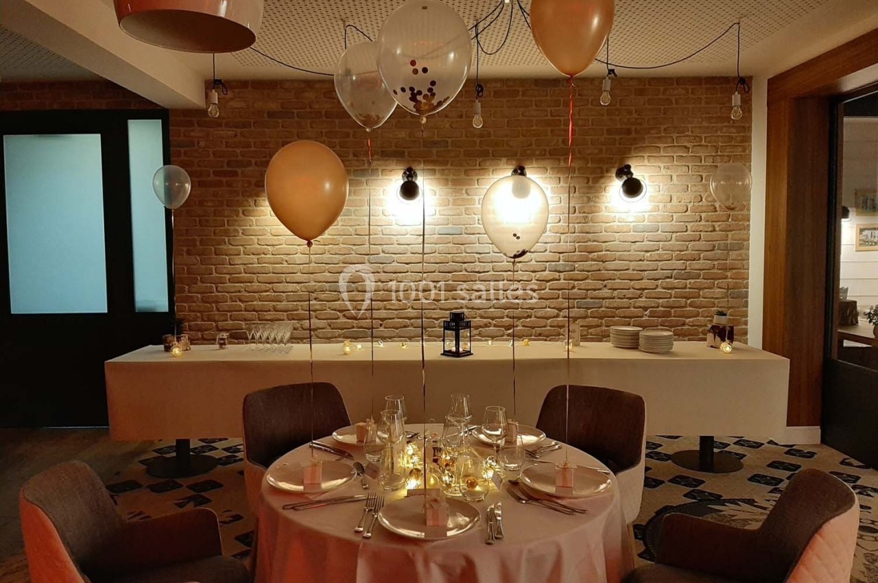 Table décorée pour un événement avec nappes blanches, vaisselle élégante et ballons suspendus dans une salle éclairée.
