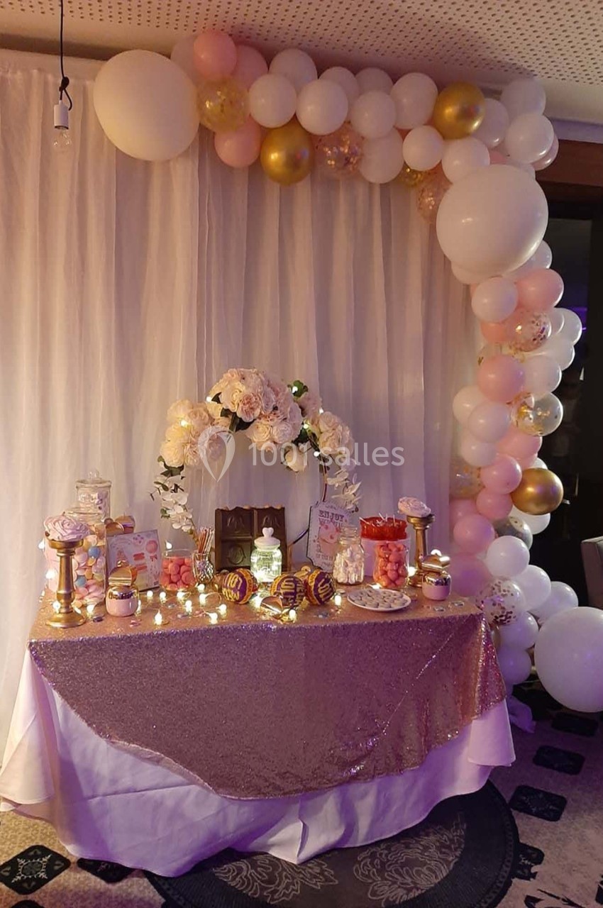 Table décorée pour une fête avec nappes scintillantes, fleurs, ballons blancs et dorés, et desserts variés.