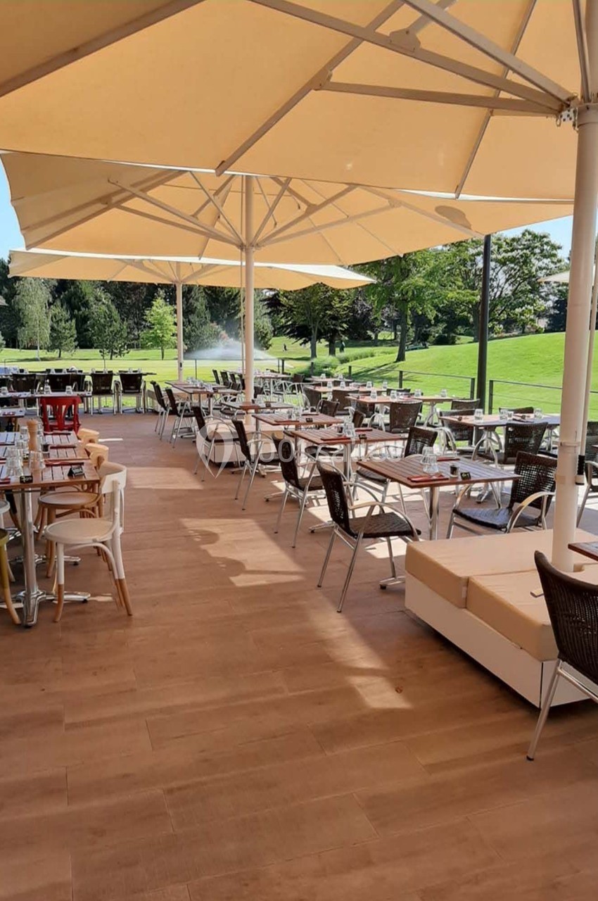 Terrasse d'un restaurant avec tables et chaises sous des parasols, donnant sur un espace vert par une journée ensoleillée.