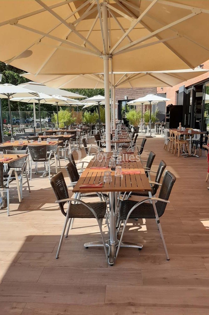 Terrasse extérieure avec tables en bois, chaises en osier et parasols, entourée de verdure et baignée de lumière naturelle.