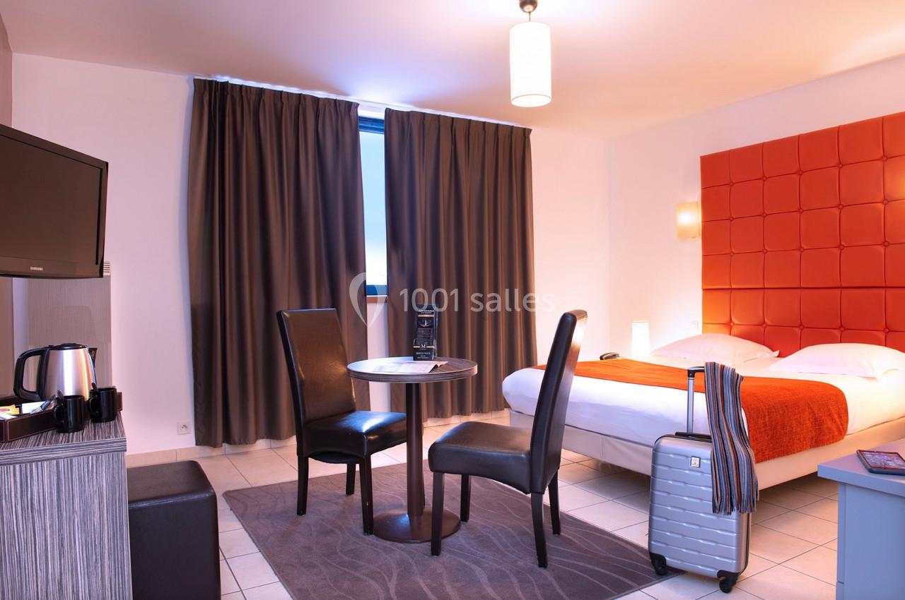 Chambre d'hôtel lumineuse avec lit double, table, chaises, valise, télévision, rideaux fermés et éclairage moderne.