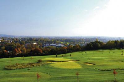 Vue d'un terrain de golf verdoyant avec des arbres et une ville en arrière-plan sous un ciel dégagé.