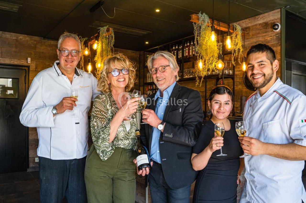 Cinq personnes souriantes trinquent avec des verres de vin dans un intérieur chaleureux décoré de plantes suspendues.