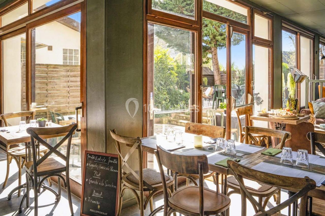 Salle de restaurant lumineuse avec grandes baies vitrées, tables en bois dressées et vue sur un espace extérieur arboré.