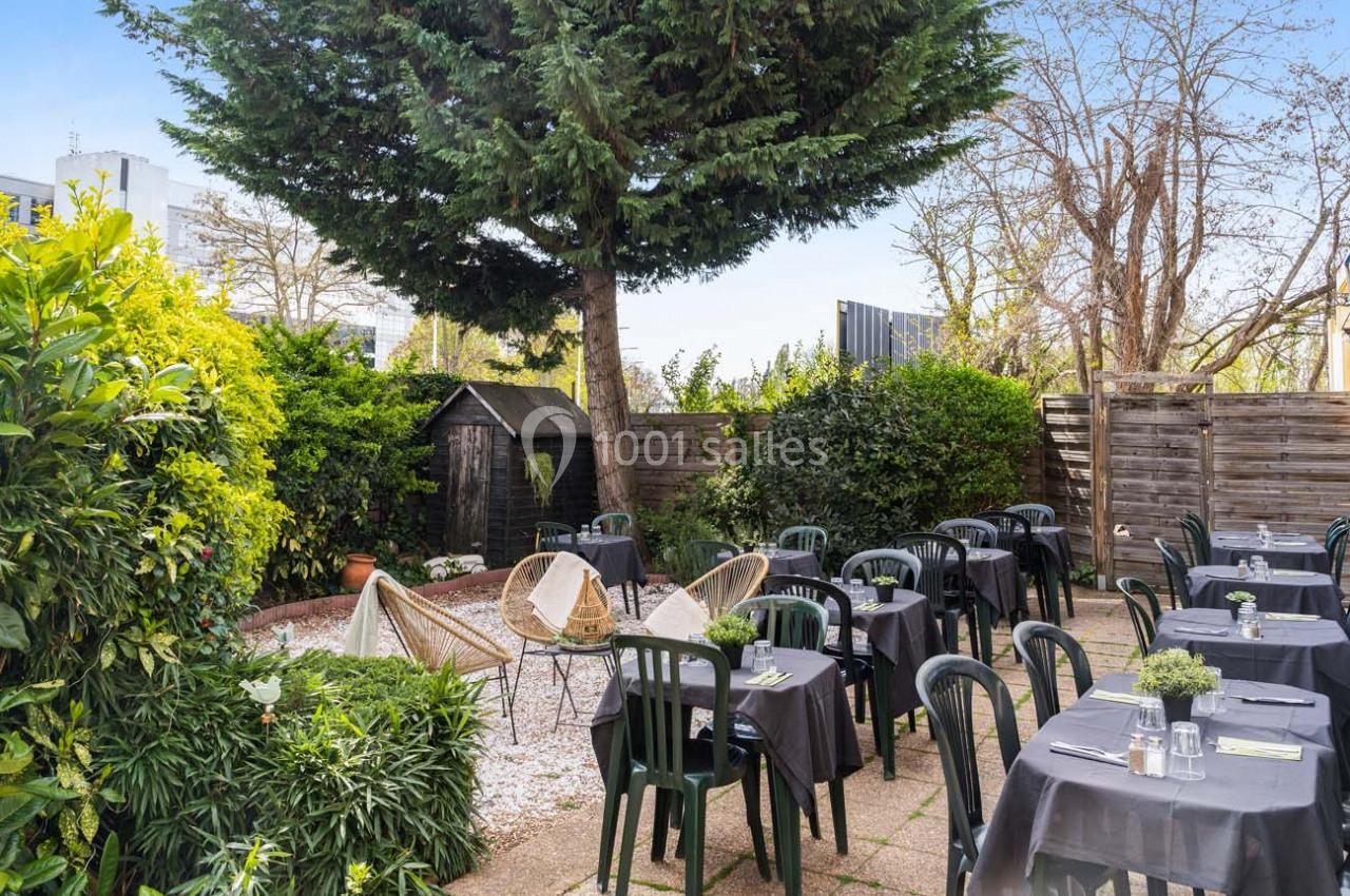 Terrasse extérieure avec tables dressées, chaises noires, végétation abondante et un grand arbre au centre.