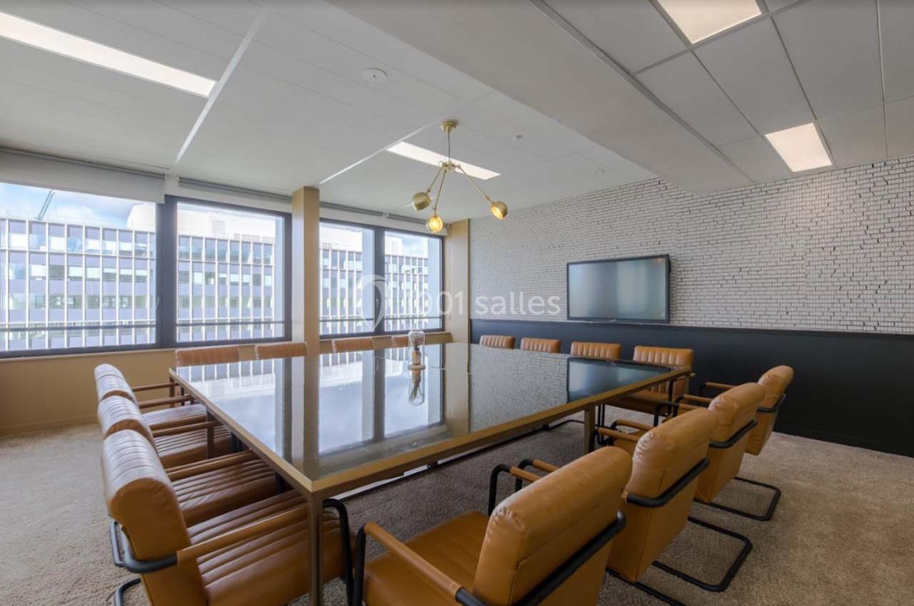 Salle de réunion lumineuse avec une grande table en verre, des chaises en cuir marron et un écran mural.