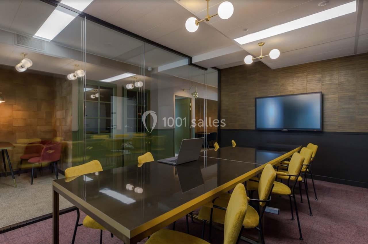 Salle de réunion moderne avec table longue, chaises jaunes, écran mural et cloison vitrée séparant un espace adjacent.