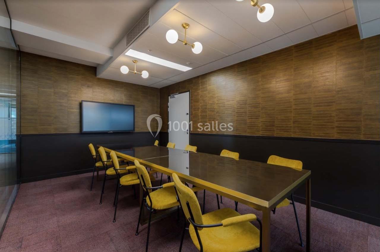 Salle de réunion avec table rectangulaire, chaises jaunes, écran mural et éclairage suspendu.
