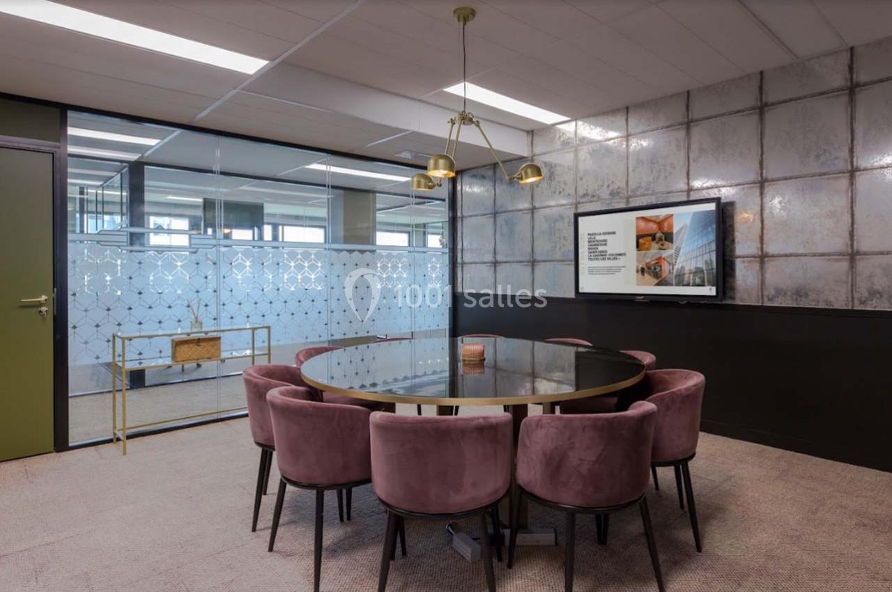 Salle de réunion moderne avec table ronde en verre, chaises roses, écran mural et cloison vitrée décorative.