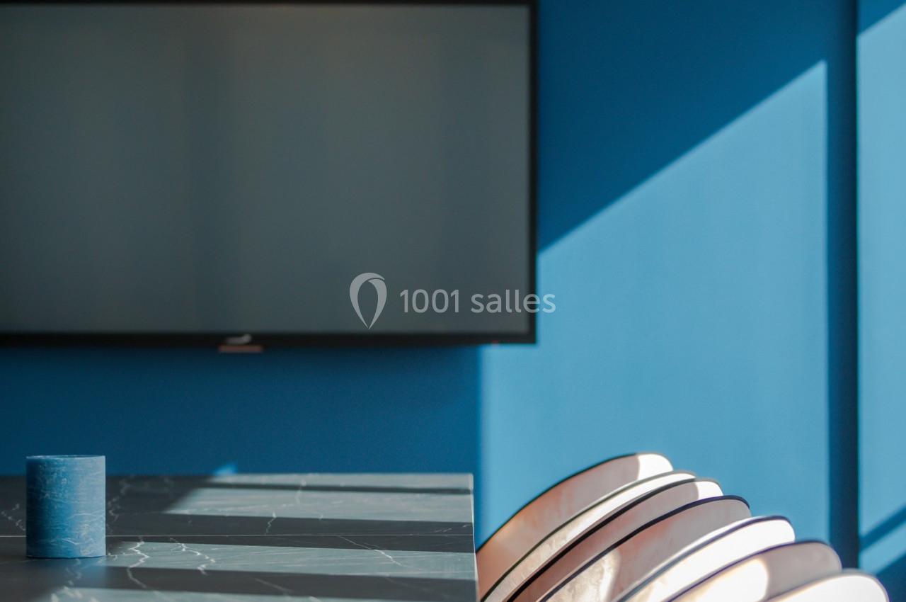 Salle de réunion avec mur bleu, table en marbre sombre, chaises claires et écran mural éteint.