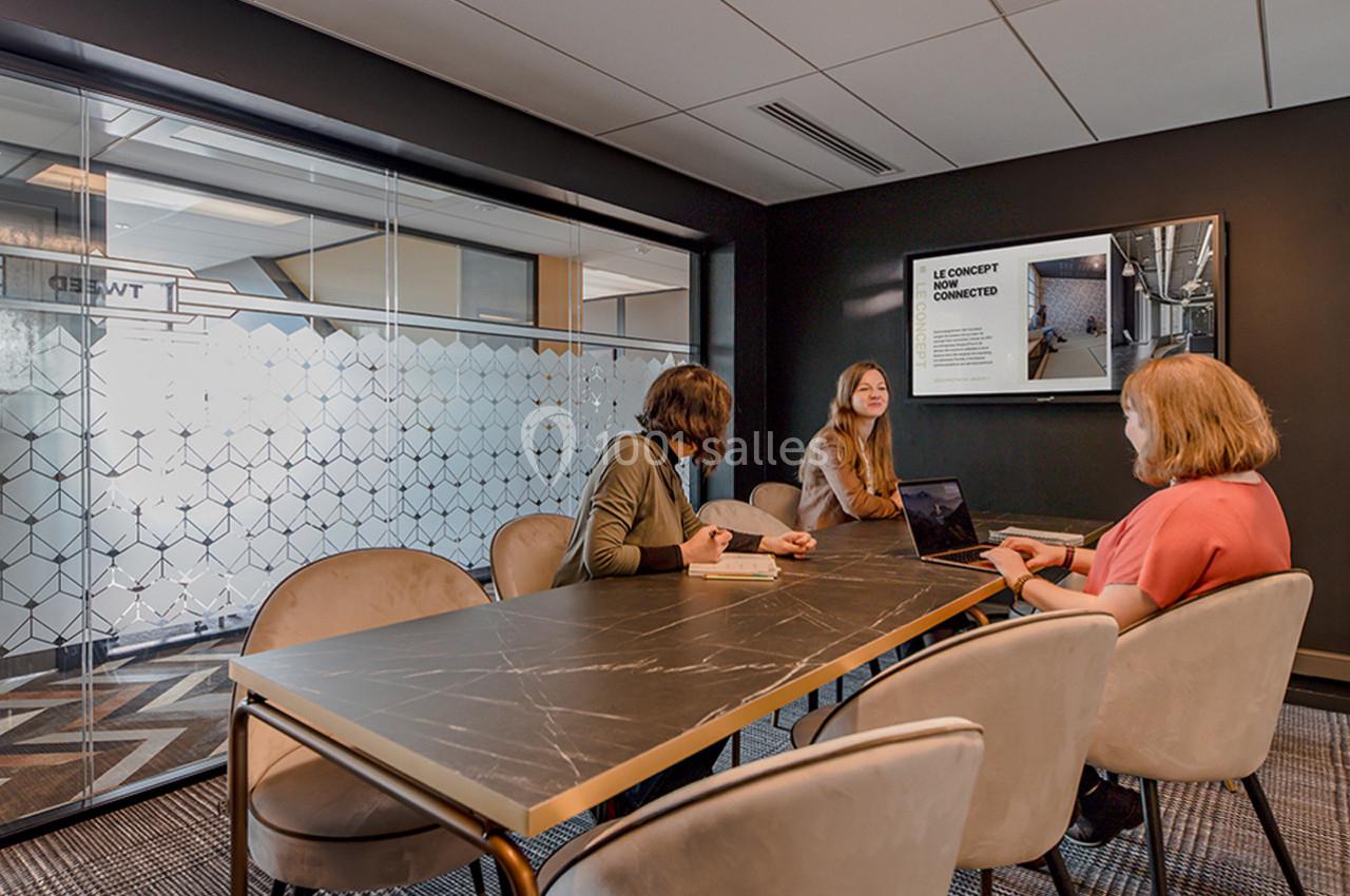 Trois personnes discutent autour d'une table dans une salle de réunion moderne avec un écran affichant une présentation.