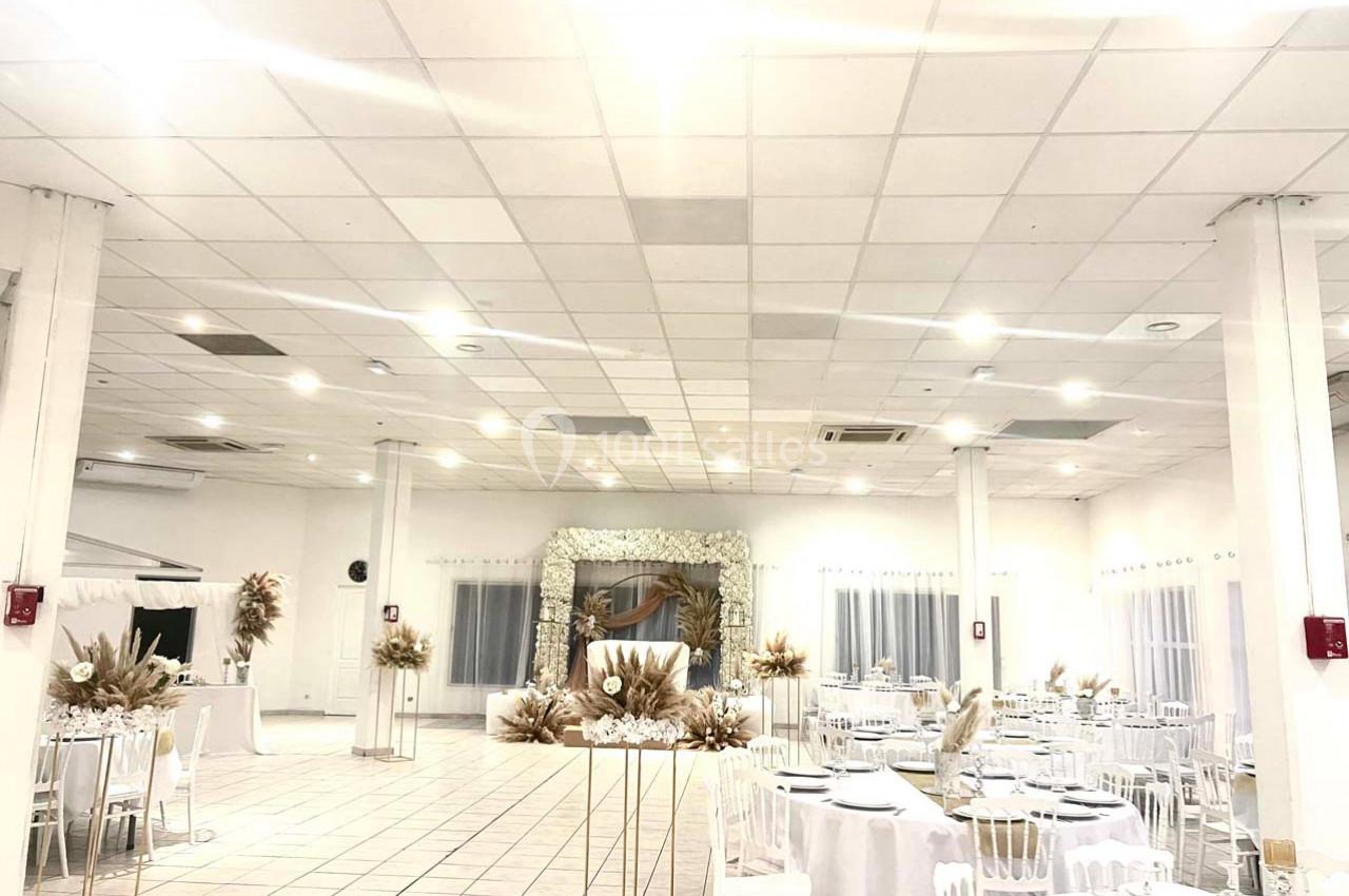 Salle de réception lumineuse avec tables dressées, décoration florale blanche et arche ornée de fleurs en arrière-plan.