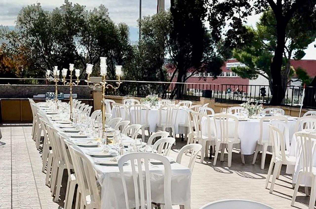 Tables dressées en extérieur avec nappes blanches, chaises en plastique et chandeliers, entourées d'arbres.