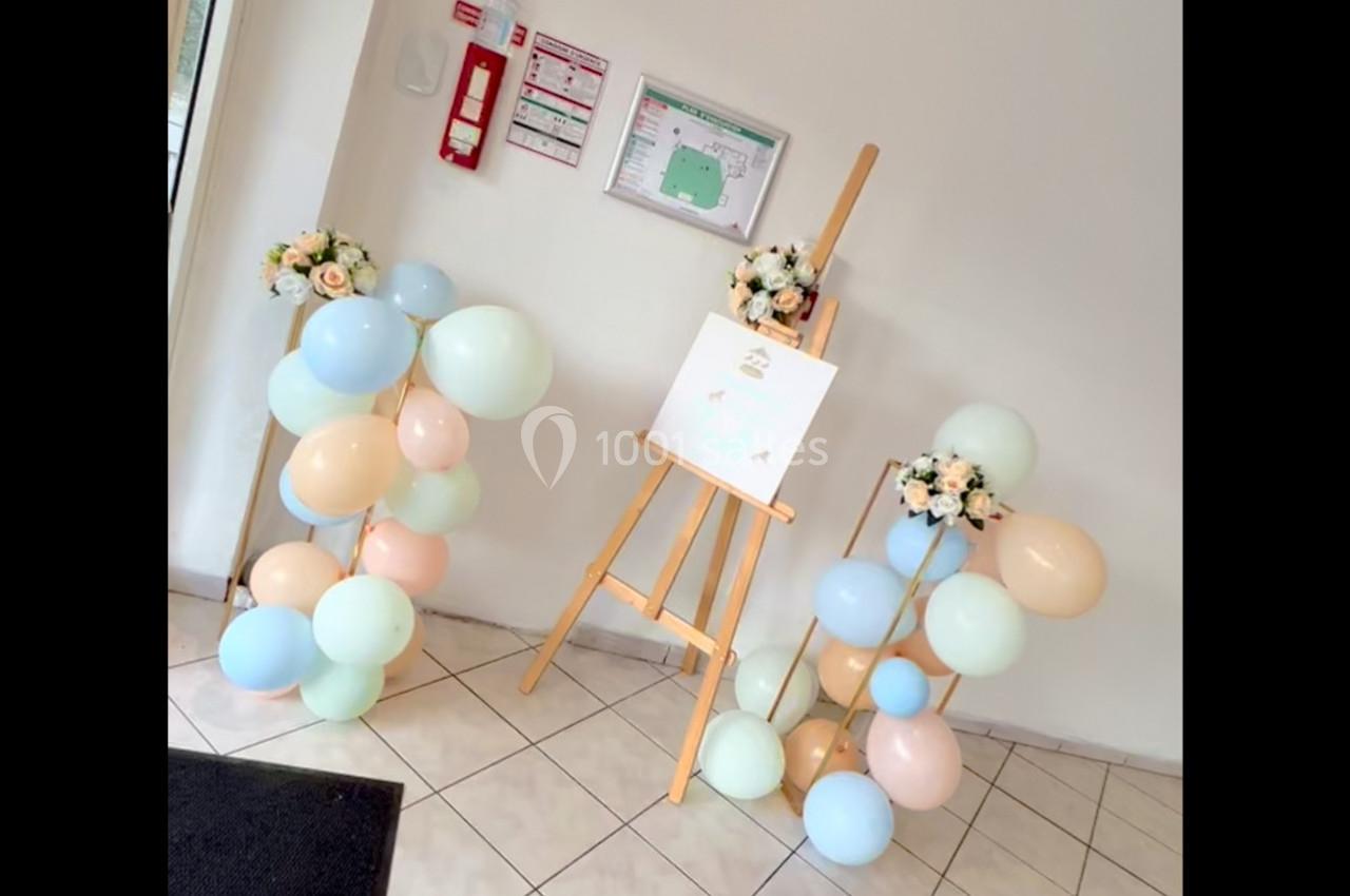 Chevalet avec tableau décoratif entouré de ballons pastel et bouquets de fleurs dans un espace intérieur lumineux.