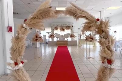 Salle de réception décorée pour un mariage, avec tables dressées, fleurs blanches et rouges, et arche florale en arrière…