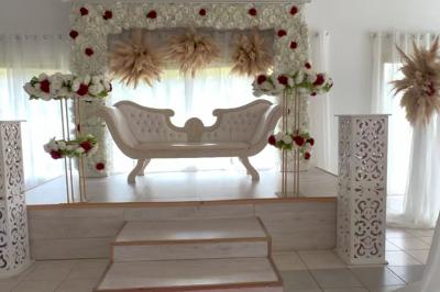 Salle de réception décorée pour un mariage, avec tables dressées, fleurs blanches et rouges, et arche florale en arrière…