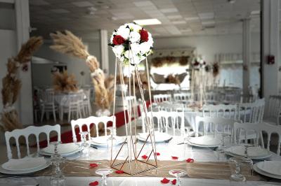 Salle de réception décorée pour un mariage, avec tables dressées, fleurs blanches et rouges, et arche florale en arrière…