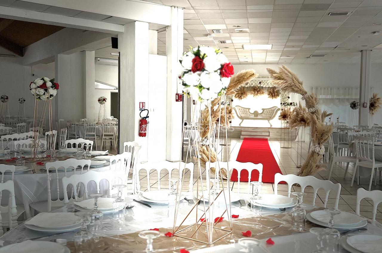Salle de réception décorée avec des tables dressées, des chaises blanches et une arche florale près d'un tapis rouge.