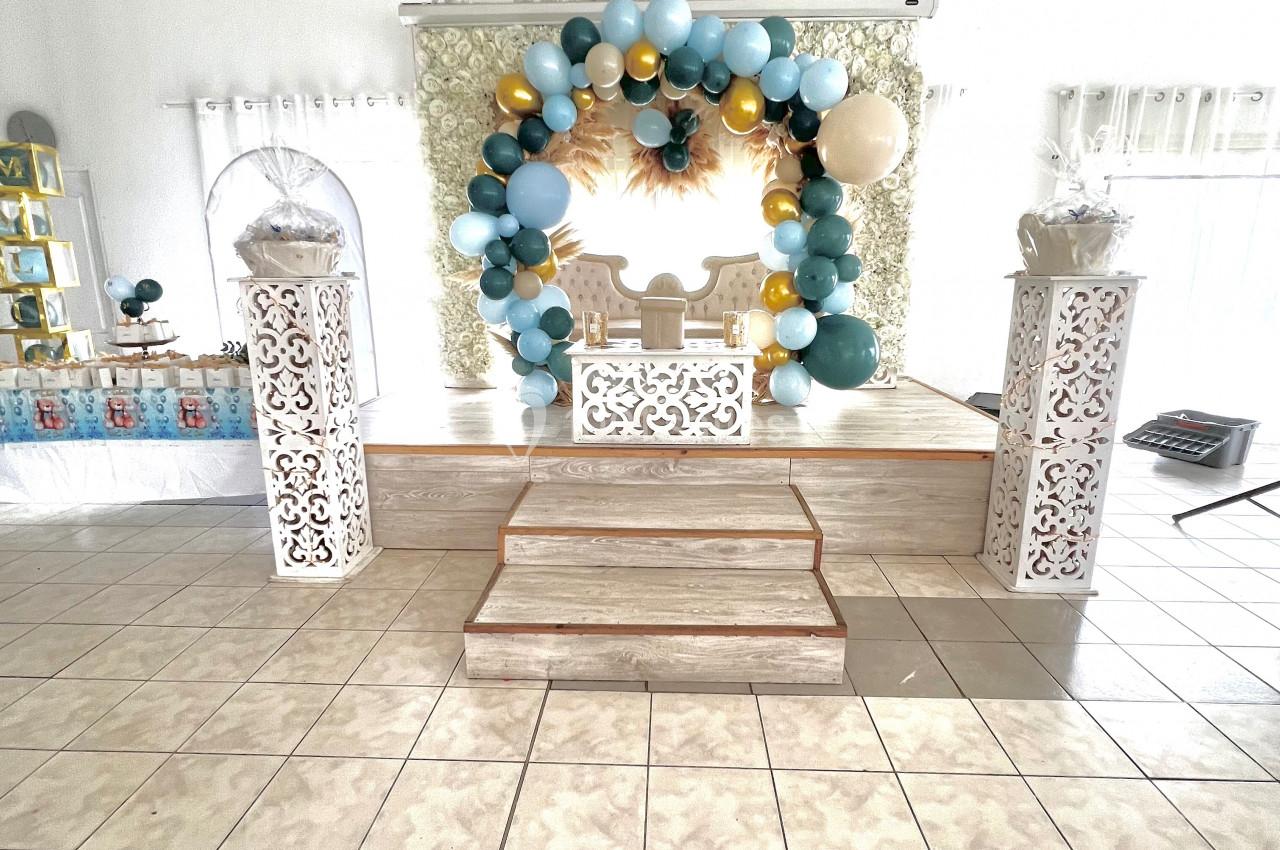 Estrade décorée avec un trône blanc, entourée d'un arc de ballons bleus, blancs et dorés dans une salle lumineuse.