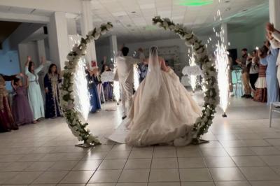 Salle de réception décorée pour un mariage, avec tables dressées, fleurs blanches et rouges, et arche florale en arrière…