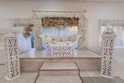 Salle de réception décorée pour un mariage, avec tables dressées, fleurs blanches et rouges, et arche florale en arrière…