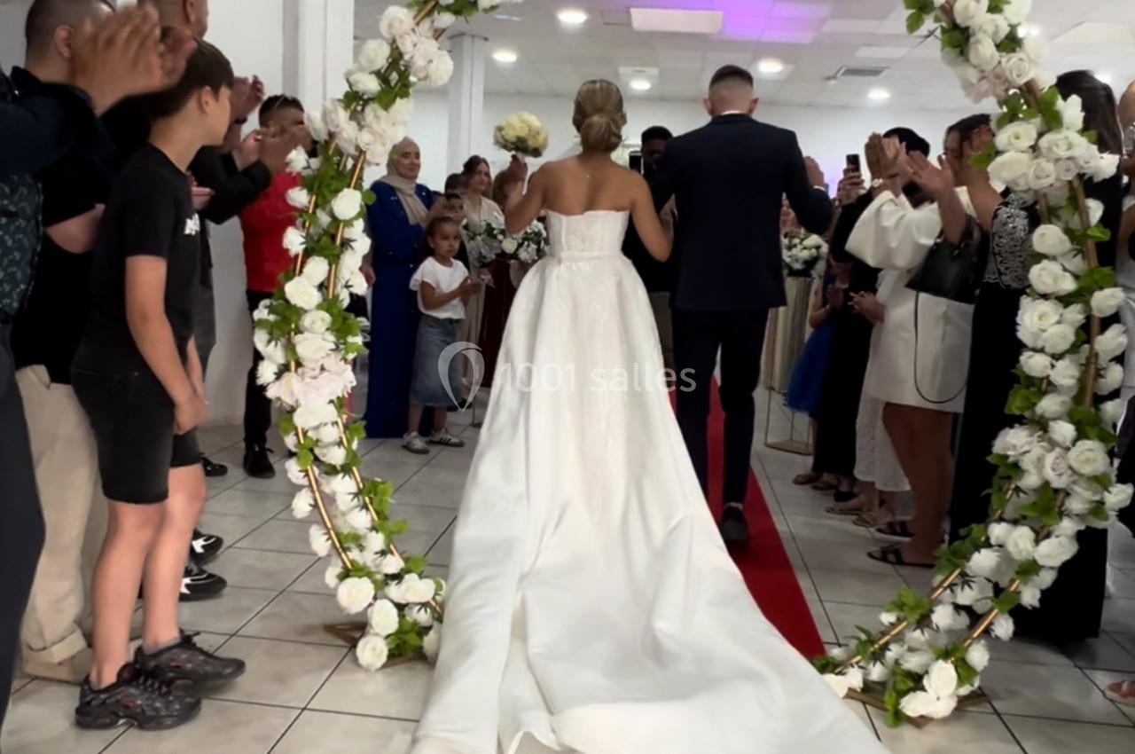Une mariée en robe blanche et son accompagnateur traversent une arche fleurie devant des invités dans une salle.